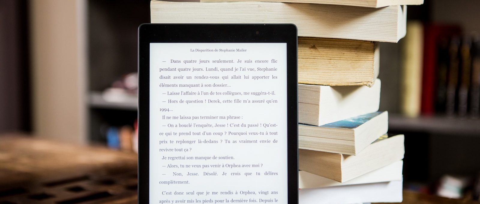 Stos położonych jedna na drugą książek na drewnianym blacie, o niego oparty włączony czytnik ebook.
