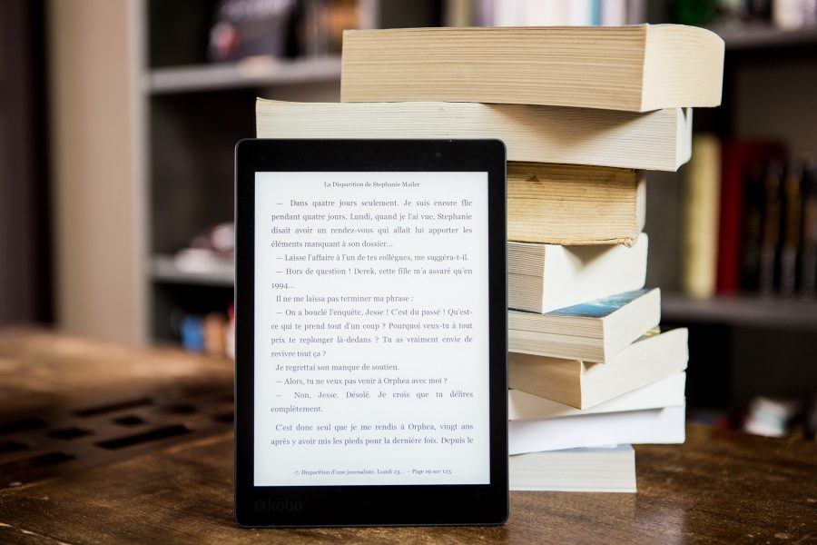 Stos położonych jedna na drugą książek na drewnianym blacie, o niego oparty włączony czytnik ebook.